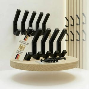 Può includere: Un set di ganci da parete in metallo nero esposti su una mensola in legno chiaro. I ganci sono angolari e moderni, con finitura opaca. Un'etichetta recita "QUALITÄT AUS DEUTSCHLAND MADE IN GERMANY". Include viti di montaggio.