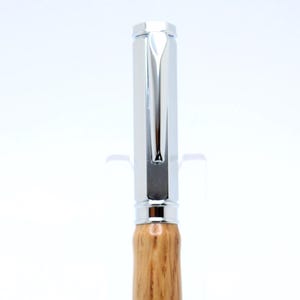 May include: A polished pen with a silver cap and accents, featuring a wooden barrel with a natural grain pattern. The pen is displayed on a clear stand against a white background. A writing instrument.