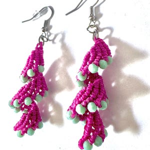 May include: A pair of dangle earrings featuring vibrant magenta macrame cord knotted into a spiral shape, accented with small, round, pale mint green beads integrated throughout the design, finished with silver-toned metal fishhook ear wires.