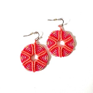 Puede incluir: Un par de pendientes circulares de macramé con un degradado vibrante de hilo rosa, coral y naranja. Cada pendiente tiene un orificio central y está construido con intrincados patrones de nudos que irradian hacia afuera, con ganchos de metal plateado.