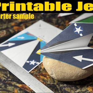 May include: A paper airplane with a navy blue and white design, featuring a star and arrow, rests on a stone. The text "Printable Jet" and "Starter sample" are displayed in yellow. A ruler and printed paper are also visible.