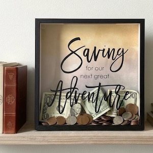 May include: A square black shadow box frame with a glass front featuring the black text Saving for our next great Adventure. The background has a soft gradient of yellow and white. Inside the frame are several US dollar bills and coins.