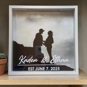 May include: White framed shadow box with a black and white silhouette of a couple. The names "Kaden & Ethan" are displayed in white script, with "EST JUNE 7, 2025" below. A small potted succulent is visible on the left.