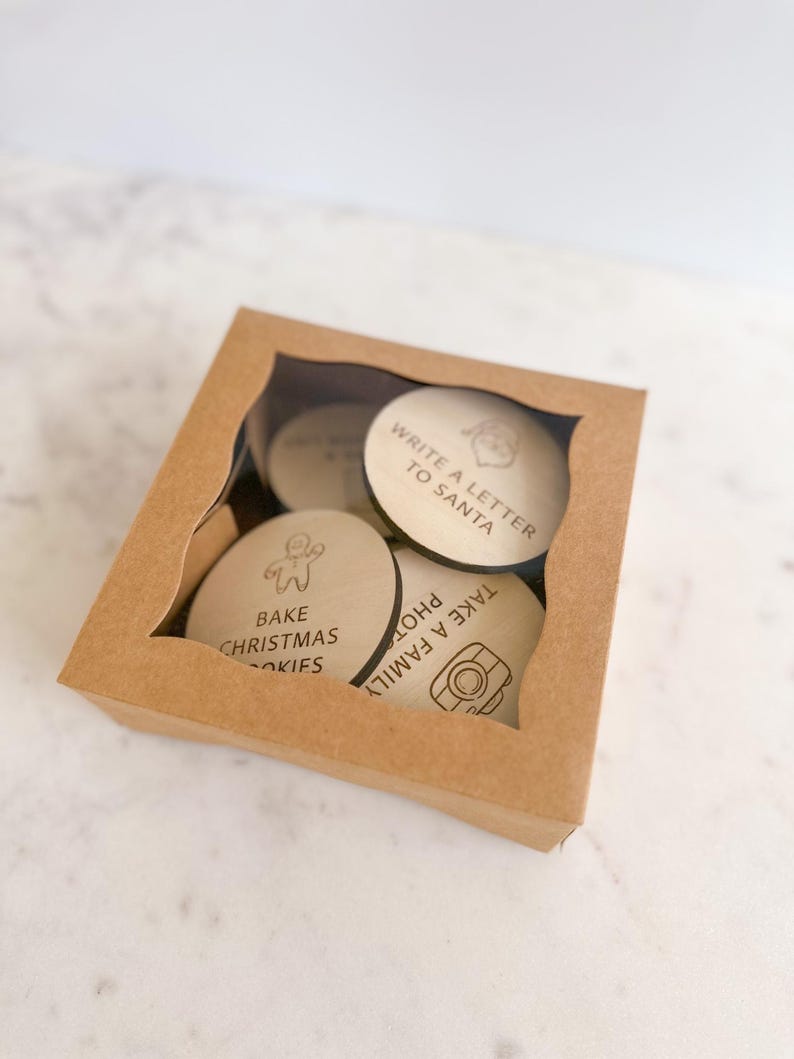 Puede incluir: Una caja de cart&oacute;n marr&oacute;n con una ventana transparente que muestra cinco c&iacute;rculos de madera con actividades navide&ntilde;as escritas en ellos. Las actividades incluyen "Escribir una carta a Santa Claus", "Hornear galletas de Navidad", "Tomar una foto familiar", "Decorar el &aacute;rbol", y "Hacer un mu&ntilde;eco de nieve".