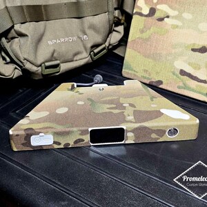Könnte beinhalten: Ein elektronisches Gerät mit Tarnmuster, einer schwarzen rechteckigen Öffnung und mehreren Knöpfen. Ein passendes Panel mit Tarnmuster und ein grüner Rucksack mit dem Text "SPARROW 16" befinden sich im Hintergrund. Das Gerät ist wahrscheinlich ein kundenspezifischer Artikel.