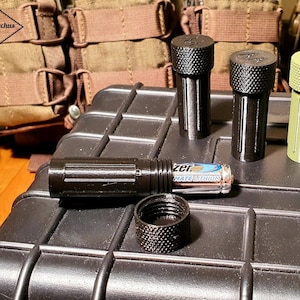 May include: A collection of small, cylindrical containers in black and olive green, with textured caps. One container is open, revealing a battery. The items are arranged on a black, textured surface, with tactical gear in the background.