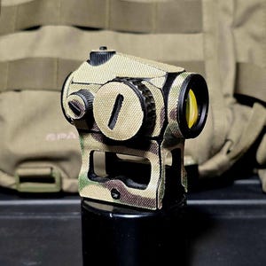 Op de afbeelding: Een camouflage-gepatroneerd rood puntvizier met een gele lens. Het vizier is gemonteerd op een zwarte basis en staat op een zwart oppervlak.