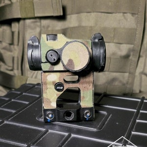 Op de afbeelding: Een zwart en groen camouflage-gepatroneerd rood puntvizier met een zwarte basis en een kleine zwarte knop aan de bovenkant. Het vizier is gemonteerd op een zwart oppervlak.