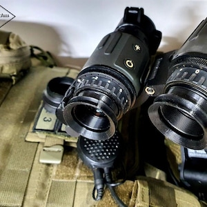 May include: Two black and gray night vision scopes with a focus ring and a dial. The scopes are mounted on a tactical vest.
