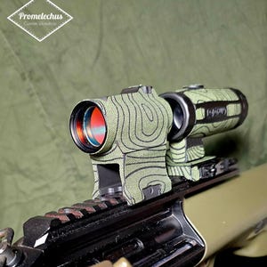 Op de afbeelding: Een groen en zwart patroon vizier gemonteerd op een geweer. De scope heeft een rode lens en is ontworpen met een topografisch kaartpatroon. Het geweer is olijfgroen en zwart. De afbeelding bevat de tekst "Promelechus Custom Workshop."