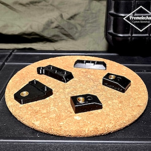 Op de afbeelding: Een assortiment zwarte, geometrische hardware-onderdelen met messingkleurige schroefdraadinzetstukken is gerangschikt op een kurkoppervlak. De onderdelen variëren in vorm en grootte, sommige met meerdere gaten. De achtergrond omvat een donker, getextureerd oppervlak en een logo.
