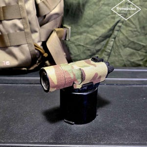 Puede incluir: Una mira óptica negra y con patrón de camuflaje, montada sobre una base cilíndrica negra. El patrón de camuflaje incluye tonos de verde, marrón y beige. El fondo incluye una tela verde y una mochila beige.