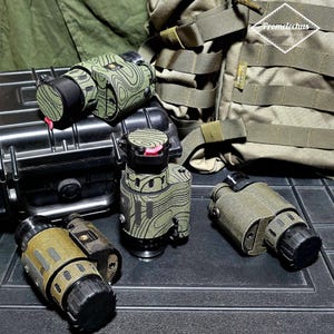 May include: Several olive green and black tactical devices with a topographic pattern are displayed on a black surface. The devices have various shapes and sizes, with black lenses and red accents. A military-style backpack and a black case are in the background.
