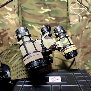 May include: Close-up of a night vision device with three lenses. The device has a black and tan camouflage pattern. The text "NOT NODS" is on a white sticker on a black case.