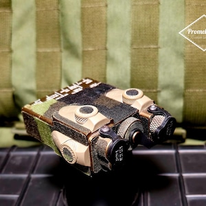 Op de afbeelding: Een tactische radio met camouflagepatroon en meerdere aanhangsels, tegen een zwarte achtergrond.