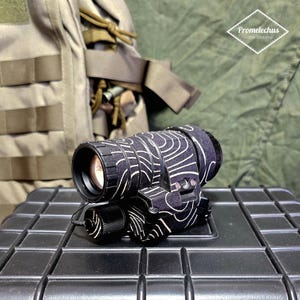 Puede incluir: Un dispositivo óptico con estampado en blanco y negro con el texto "Promelechus Custom Workshop". El dispositivo está sobre una superficie negra, con un chaleco táctico color canela en el fondo.