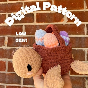 May include: A crocheted turtle carrying a basket of colorful crocheted eggs. The turtle is brown and has a cute face. The basket is brown and has a handle. The eggs are pink, blue, and purple. The text "Digital Pattern" and "Low Sew!" is written on the image.