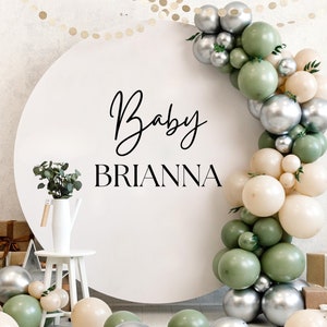 May include: A white round backdrop with the text "Baby Brianna" in black calligraphy. A garland of green, cream, and silver balloons is draped around the backdrop.