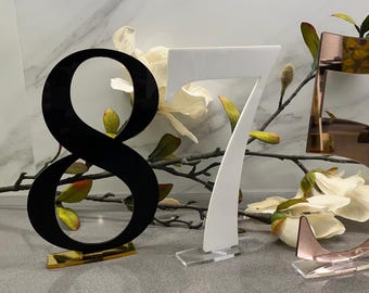 Könnte beinhalten: Drei Acryl-Tischnummern, 8, 7 und 5, in Schwarz, Weiß und Roségold. Die Nummern stehen auf klaren Acrylständern.