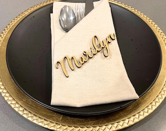 May include: A place setting featuring a black plate with a gold rim, topped with a folded beige napkin. Silverware, including a spoon, fork, and knife, is tucked into the napkin. A wooden name tag that says "Marilyn" is placed on the napkin.