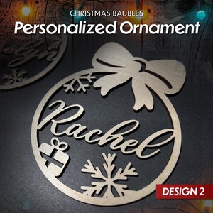 May include: A wooden Christmas bauble in the shape of a circle with a bow at the top. The bauble is decorated with snowflakes, a gift box, and the name "Rachel". The bauble is labelled "DESIGN 2".