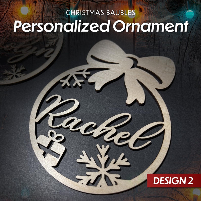 May include: A wooden Christmas bauble in the shape of a circle with a bow at the top. The bauble is decorated with snowflakes, a gift box, and the name "Rachel". The bauble is labelled "DESIGN 2".