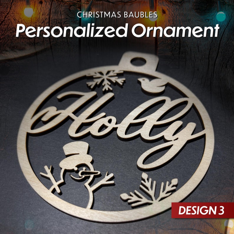 May include: A wooden Christmas bauble with the word "Holly" cut out in a decorative font. The bauble also features a snowman, snowflakes, and a bird. The bauble is labelled "Design 3".