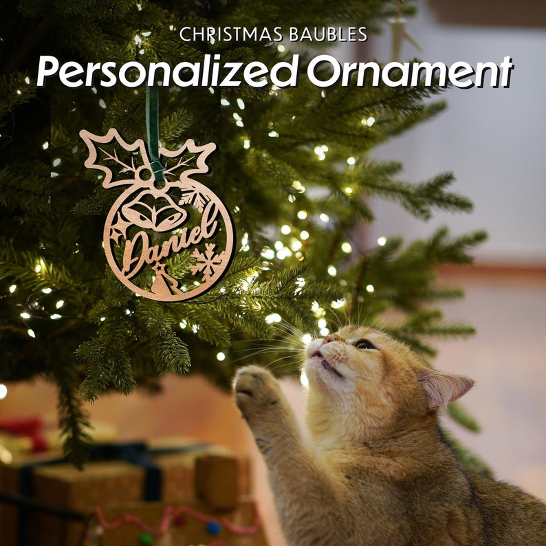 May include: A wooden Christmas bauble shaped like a ball with the name "Daniel" inscribed on it. The bauble is hanging on a Christmas tree with a cat looking up at it.