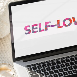 May include: A laptop displays the words "SELF-LOVE" in colorful, patterned letters. The laptop sits on a textured, off-white surface, with a glass of coffee visible in the foreground. The keyboard is in focus.