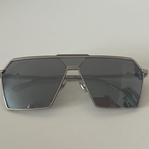 Puede incluir: Gafas de sol con montura plateada y lentes gris oscuro. Las gafas tienen un diseño geométrico con doble puente y detalles decorativos en las patillas. Las lentes son rectangulares y la montura es metálica.