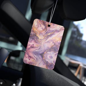 以下が含まれることがあります： 紫、ピンク、金色のマーブル模様の長方形の車用芳香剤。白い紐で吊り下げられており、背景には車の内装が写っています。