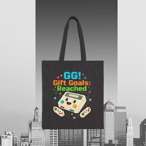 Puede incluir: Bolso tote negro con un gráfico colorido de una consola de videojuegos retro y mandos, con el texto "GG! Gift Goals: Reached." El diseño incluye estrellas y corazones. El bolso tiene dos asas negras.