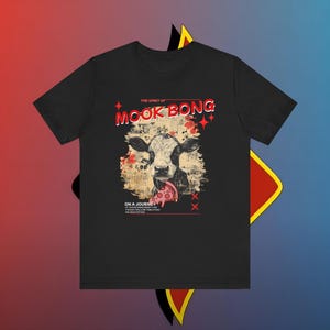 Puede incluir: Camiseta negra con un gráfico de una vaca comiendo pizza. El texto "MOOK BONG" está encima de la vaca, con las palabras "ON A JOURNEY" debajo. Acentos y estrellas rojas completan el diseño.