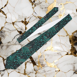 May include: Two neckties with a circuit board design. The ties are black with teal and white accents, and the design resembles the internal components of a computer. The ties are displayed on a white and gold marble surface.