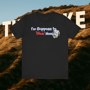 Puede incluir: Camiseta negra con el texto "I'm Suppose to 'Dink' Now" en azul y rojo, acompañada de un rostro en escala de grises. El fondo muestra una ladera con el letrero de Hollywood.