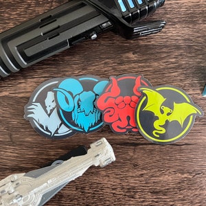 May include: Four round stickers with different fantasy creature designs. The stickers feature a blue ram, a red tentacled creature, a yellow dragon, and a white yeti. The stickers are on a wooden surface.