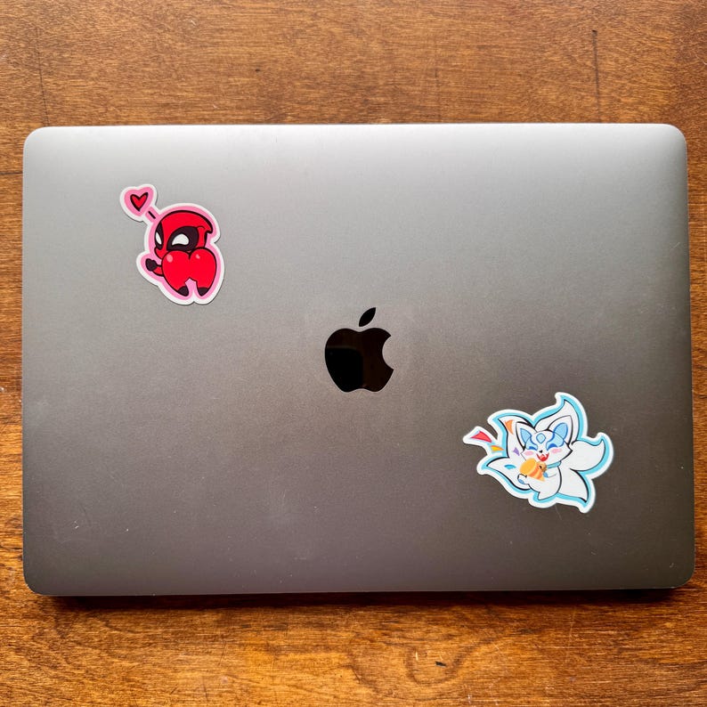 May include: A gray laptop with two stickers. One sticker is a red cartoon character with a heart, and the other is a white and blue cartoon fox-like creature.