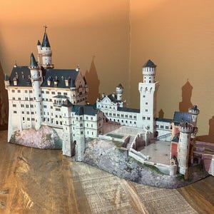 Può includere: Un modello dettagliato del castello di Neuschwanstein, con pareti bianche, tetti grigio scuro e molteplici torri. Il modello poggia su una base rocciosa ed è esposto su una superficie di legno.