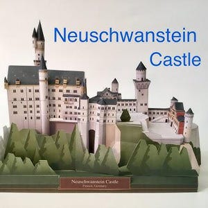 K&ouml;nnte beinhalten: Ein Papiermodell von Schloss Neuschwanstein mit wei&szlig;en W&auml;nden, grauen D&auml;chern und mehreren T&uuml;rmen. Die Basis ist von gr&uuml;nen Papierb&auml;umen umgeben. Der Text "Neuschwanstein Castle" ist rechts in Blau und auf einem braunen Etikett an der Basis gedruckt.
