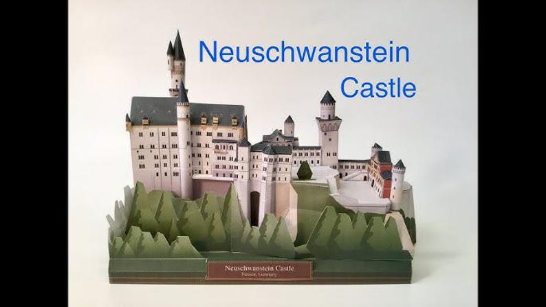 K&ouml;nnte beinhalten: Ein Papiermodell von Schloss Neuschwanstein mit wei&szlig;en W&auml;nden, grauen D&auml;chern und mehreren T&uuml;rmen. Die Basis ist von gr&uuml;nen Papierb&auml;umen umgeben. Der Text "Neuschwanstein Castle" ist rechts in Blau und auf einem braunen Etikett an der Basis gedruckt.