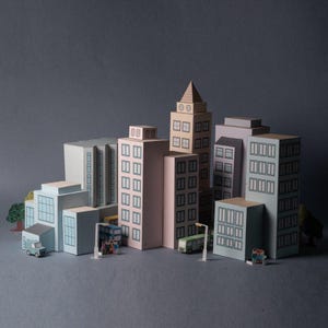 Può includere: Un modello di città in miniatura con vari edifici dai colori pastello, tra cui grattacieli e strutture più piccole. La scena comprende minuscoli veicoli, alberi e figure in miniatura, il tutto su uno sfondo grigio. Gli edifici hanno finestre dettagliate.