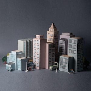 Könnte beinhalten: Ein Miniatur-Papiermodell einer Stadt mit verschiedenen Gebäuden in Pastellfarben, darunter Wolkenkratzer und kleinere Strukturen. Die Szene umfasst winzige Fahrzeuge, Bäume und Miniaturfiguren, alles vor einem grauen Hintergrund. Die Gebäude haben detaillierte Fenster.