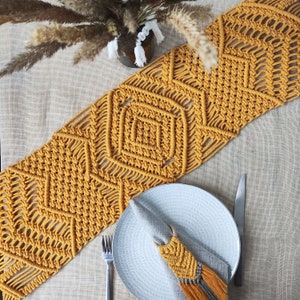 Könnte beinhalten: Senfgelber Makramee-Tischläufer mit geometrischen Rautenmustern. Ein weißer Teller, eine Gabel und ein Messer liegen auf einer beigefarbenen Tischdecke. Ein passender Serviettenring und eine Vase mit Trockenblumen vervollständigen die Tischdekoration.