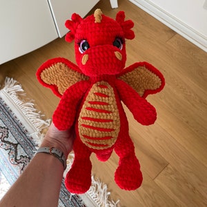 May include: A handmade red crochet dragon plush toy with tan accents on the wings and belly. The dragon has large, expressive eyes with eyelashes and a small horn. The plush is soft and cuddly, perfect for a child's toy or nursery decor.