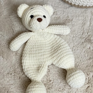May include: A cream-coloured, handmade crochet bear-shaped baby comforter. The bear has small, round ears, a brown nose, and black eyes. The body is a flat, textured design with rounded arms and legs. The comforter is on a soft, textured surface.