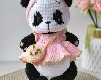 Può includere: Un giocattolo panda fatto all'uncinetto con corpo bianco e nero, che indossa un vestito rosa chiaro con orlo arricciato e una fascia abbinata con un piccolo fiocco. Il panda porta una piccola borsa rotonda rosa decorata con tre rose in tessuto.