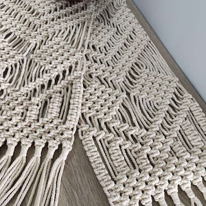 May include: Cream-colored macrame table runner with a geometric pattern and fringe detail. A wooden bowl filled with red apples sits on the runner, adding a touch of color and texture to the neutral tones.