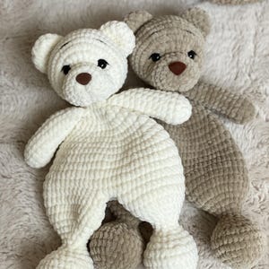 May include: Two handmade crochet teddy bears, one ivory and one tan. Both have brown button noses and black eyes. The bears are on a soft, textured surface. The ivory bear is sitting, the tan bear is lying down. A crochet basket is in the background.