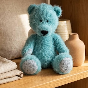 May include: A plush, turquoise teddy bear with a black nose and gray paw pads sits on a wooden shelf. The bear has a fluffy texture and is surrounded by neutral-toned decorative items, including a pillow and vases. The overall aesthetic is soft and inviting.