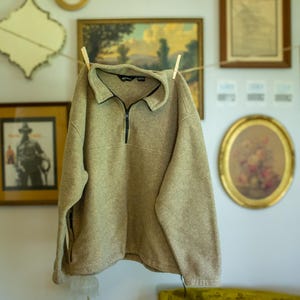 May include: A tan fleece pullover with a quarter-zip collar hangs from a clothesline. The long-sleeved top has a black zipper and collar trim. The background features framed artwork and decorative items.